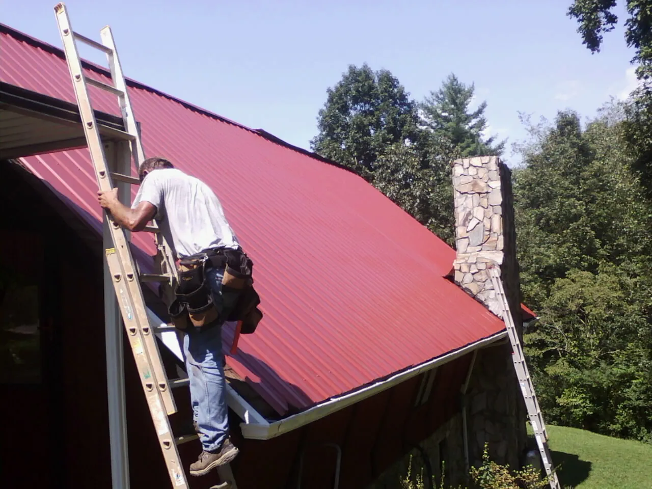 Roofer on a ladder beside a red metal roof and stone chimney — Soffit & Fascia Repair by Roofing Brothers in Hornell