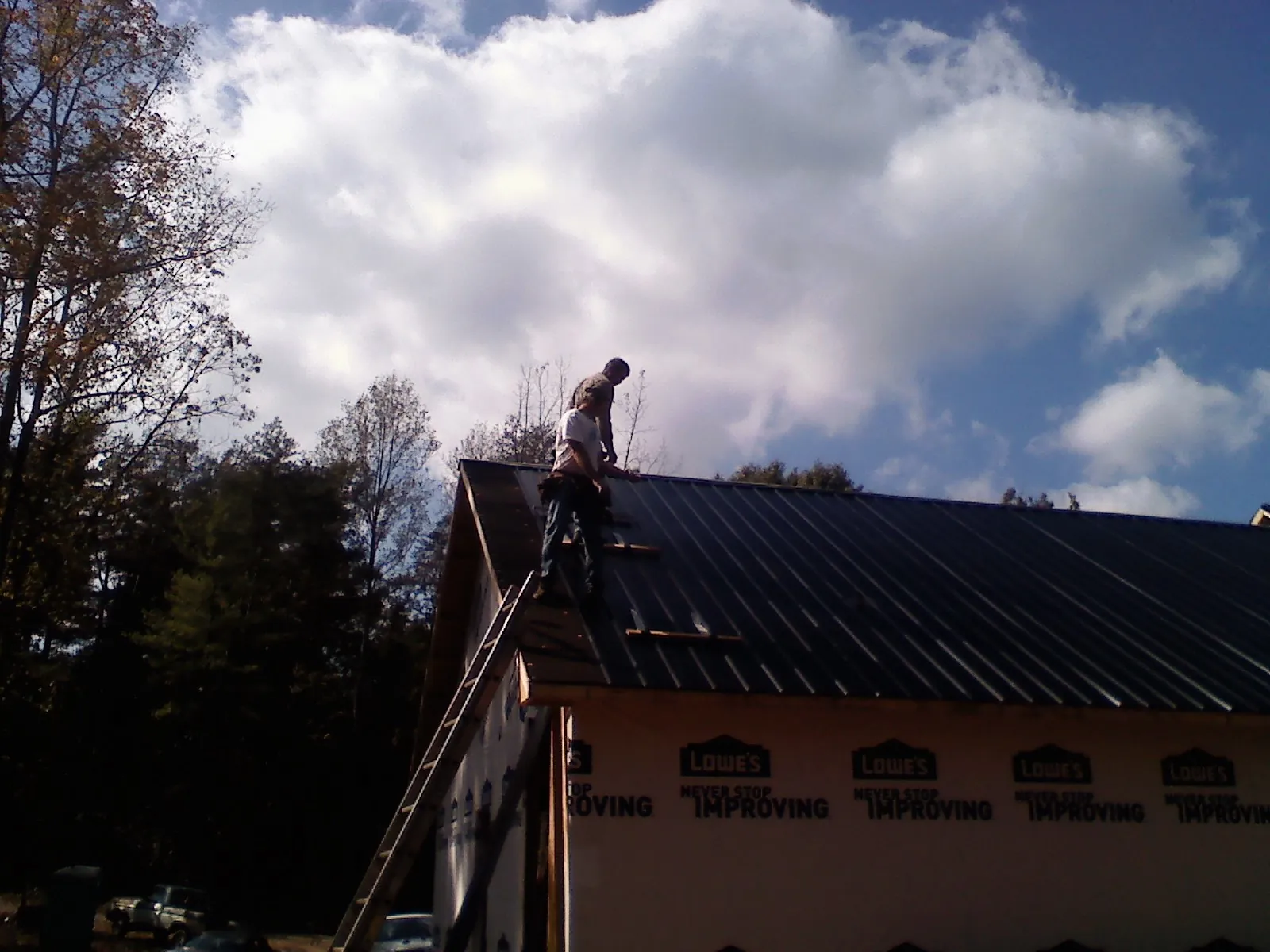 Roofer fastening dark metal panels high against a cloudy sky, from a Roof Inspection job in Hornell